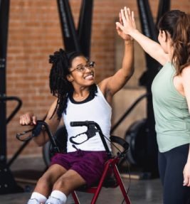The young adult woman sitting in her wheelchair gives her mid adult female friend a high five after exercising together at the gym.